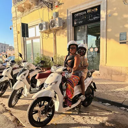 A woman and child wearing helmets sit on a white scooter in front of a rental office in Italy.