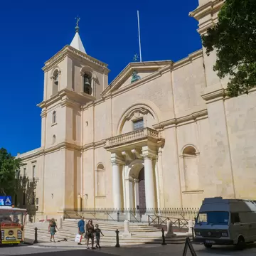 A pale limestone church facade with a bell tower and arched portico on a sunlit street.