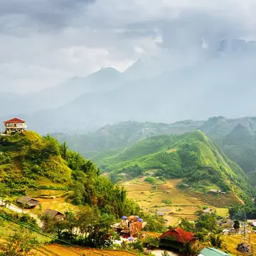 A lone building on a green hill above terraced fields and village roofs, with misty mountain ridges under heavy clouds.