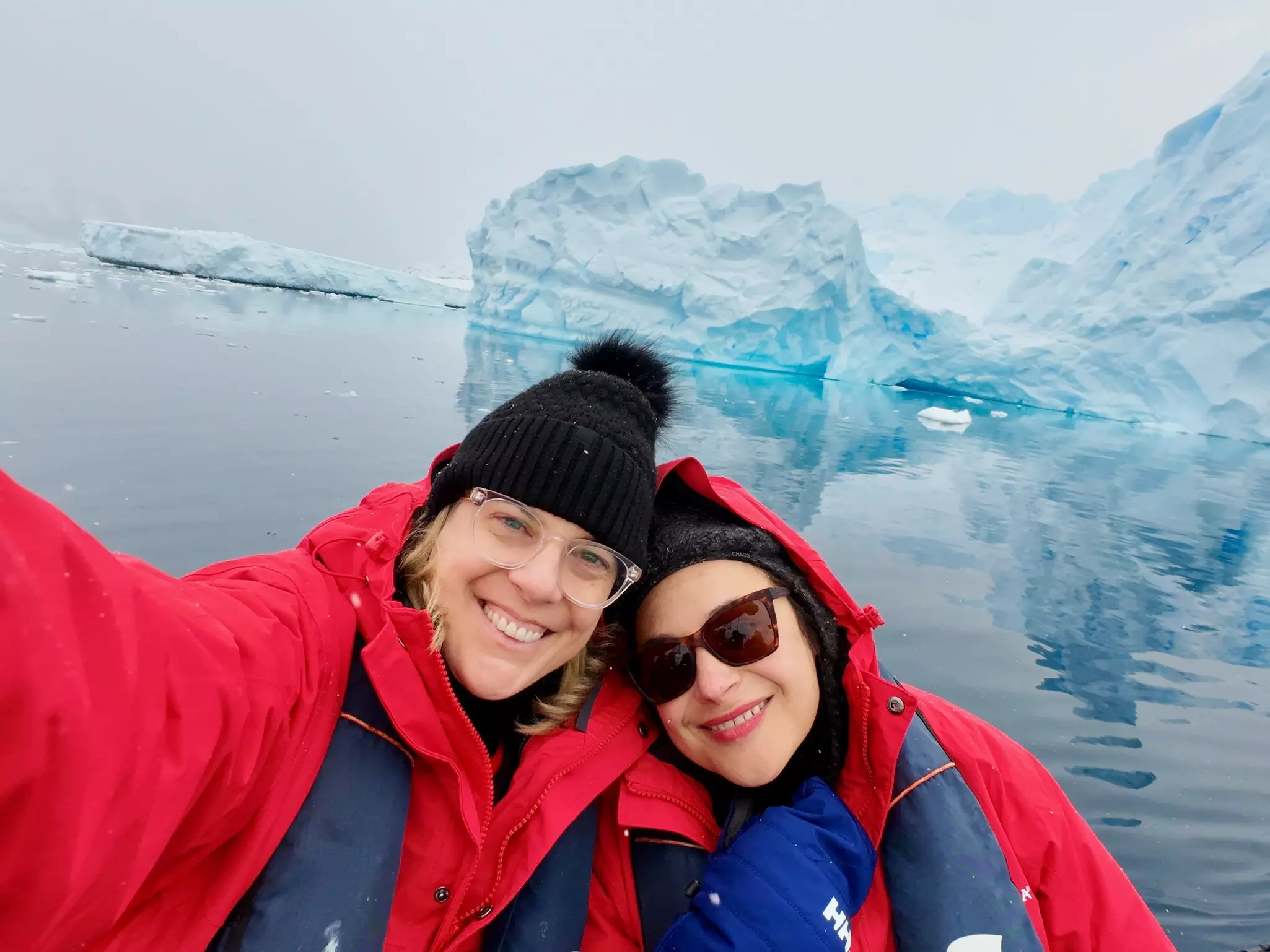 Two women pose for a selfie in front of a blue-white iceberg. They're dressed in hats and heavy coats for icy polar conditions.