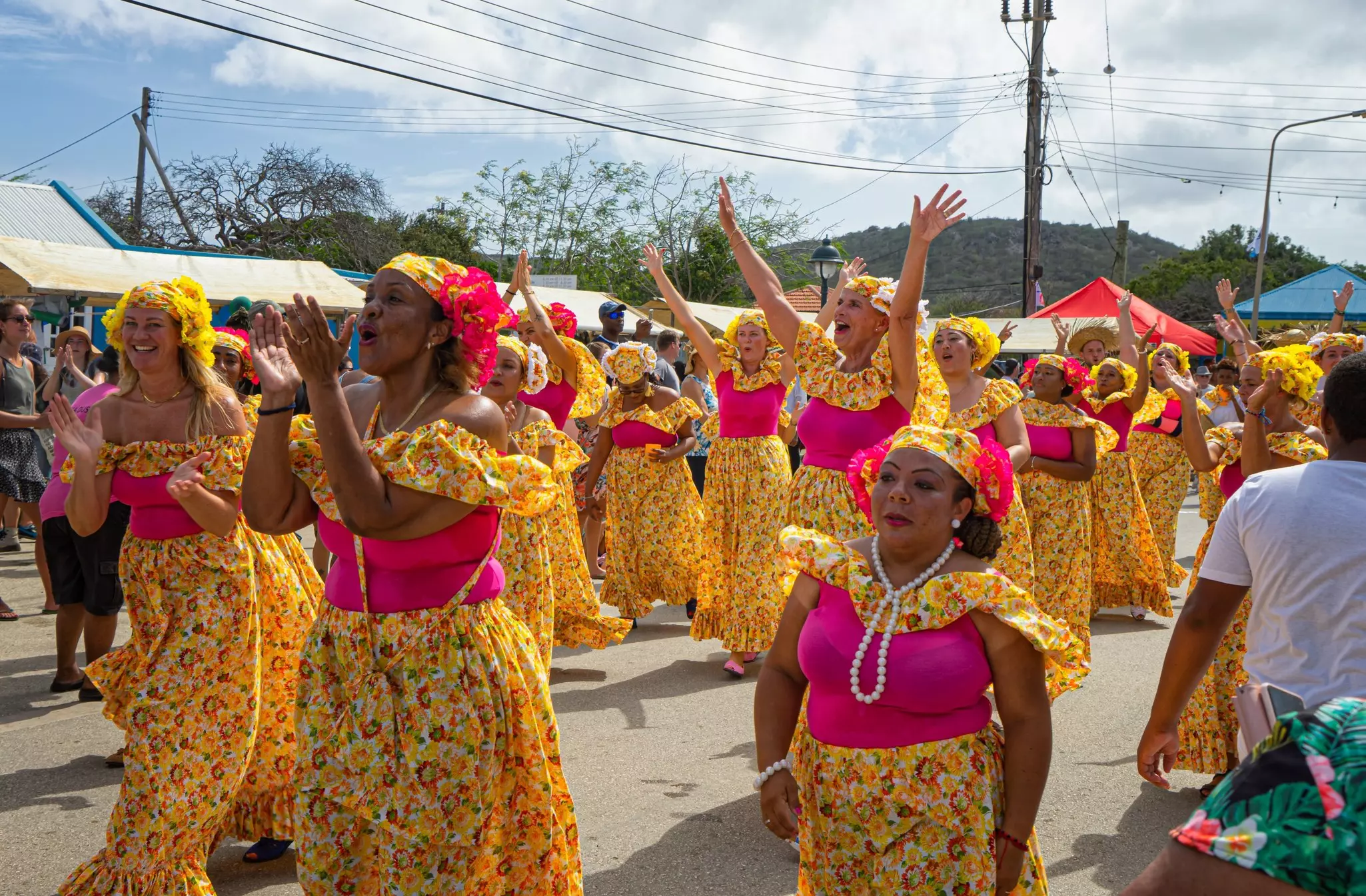 Women in matching outfits of hot pink tops and patterned yellow skirts and scarves march in a parade in a small town.