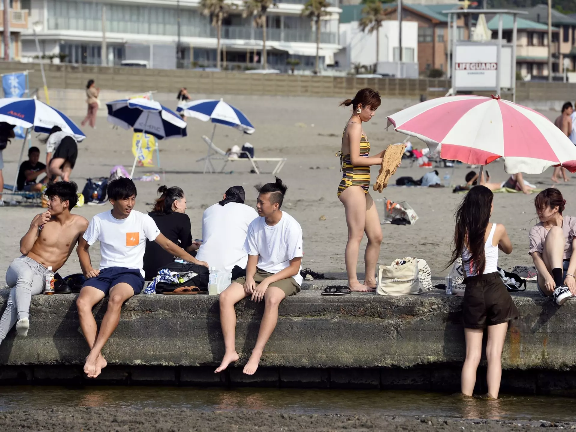 People sit on a wall at a beach near a city.