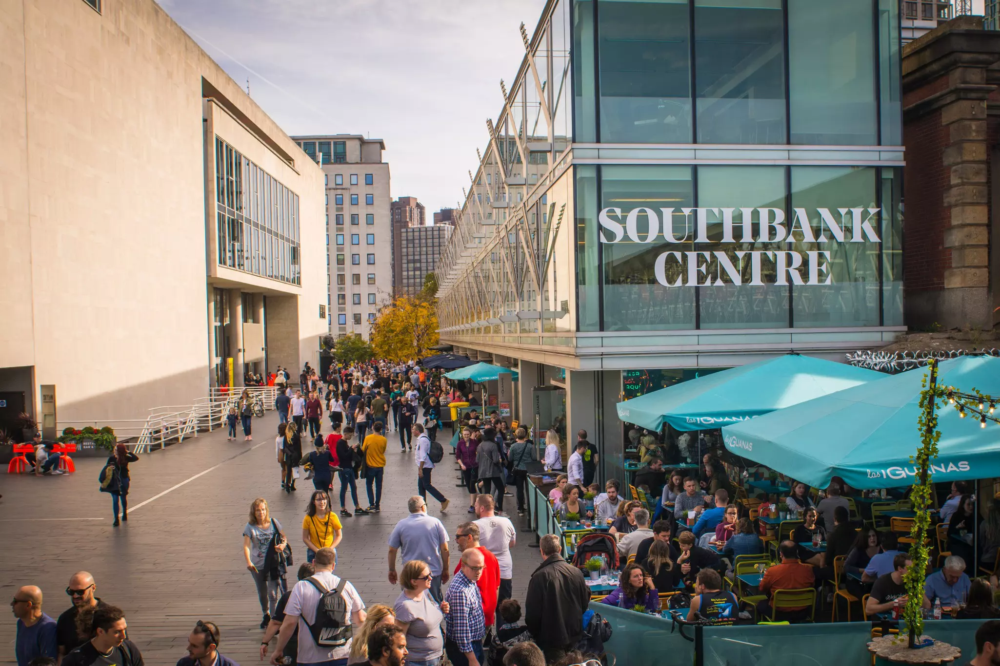 People walk between buildings of a cultural center near diners who are sat at tables outside a restaurant.