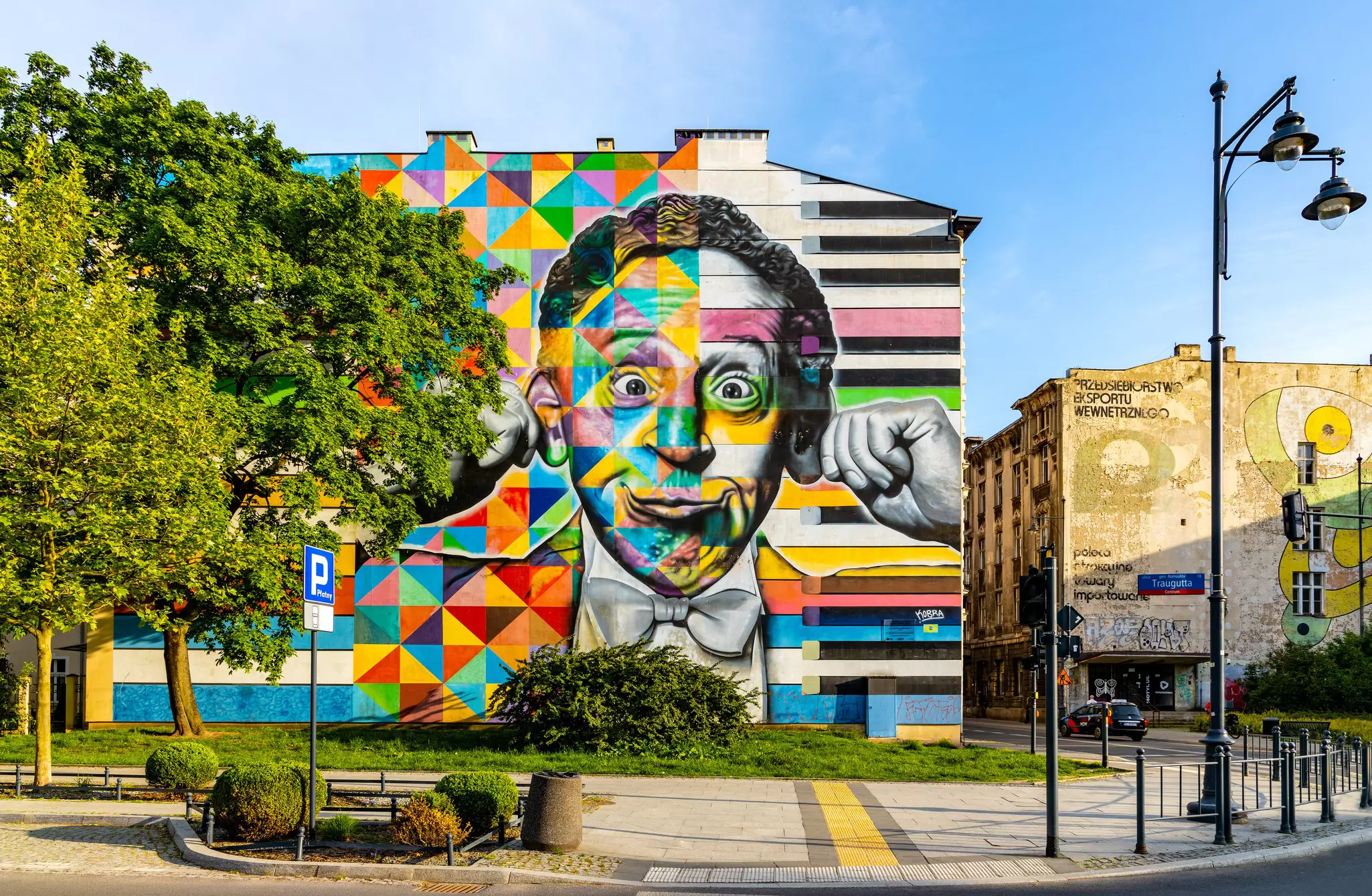 A colorful mural of a man in a bowtie making a face is painted in the side of an apartment building in a city.