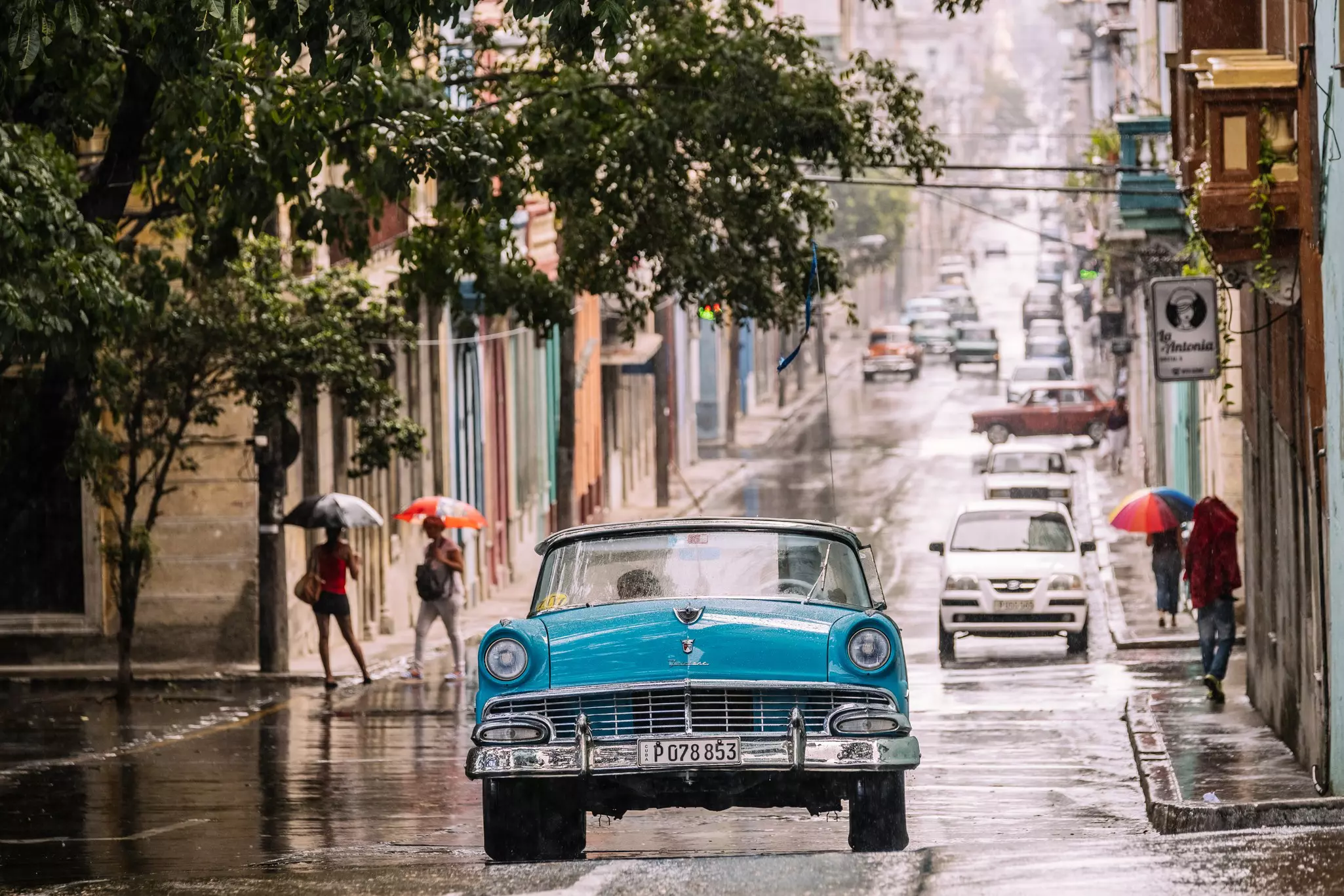 A large 1950s-style car drives along a city street in the rain. Nearby pedestrians carry umbrellas.