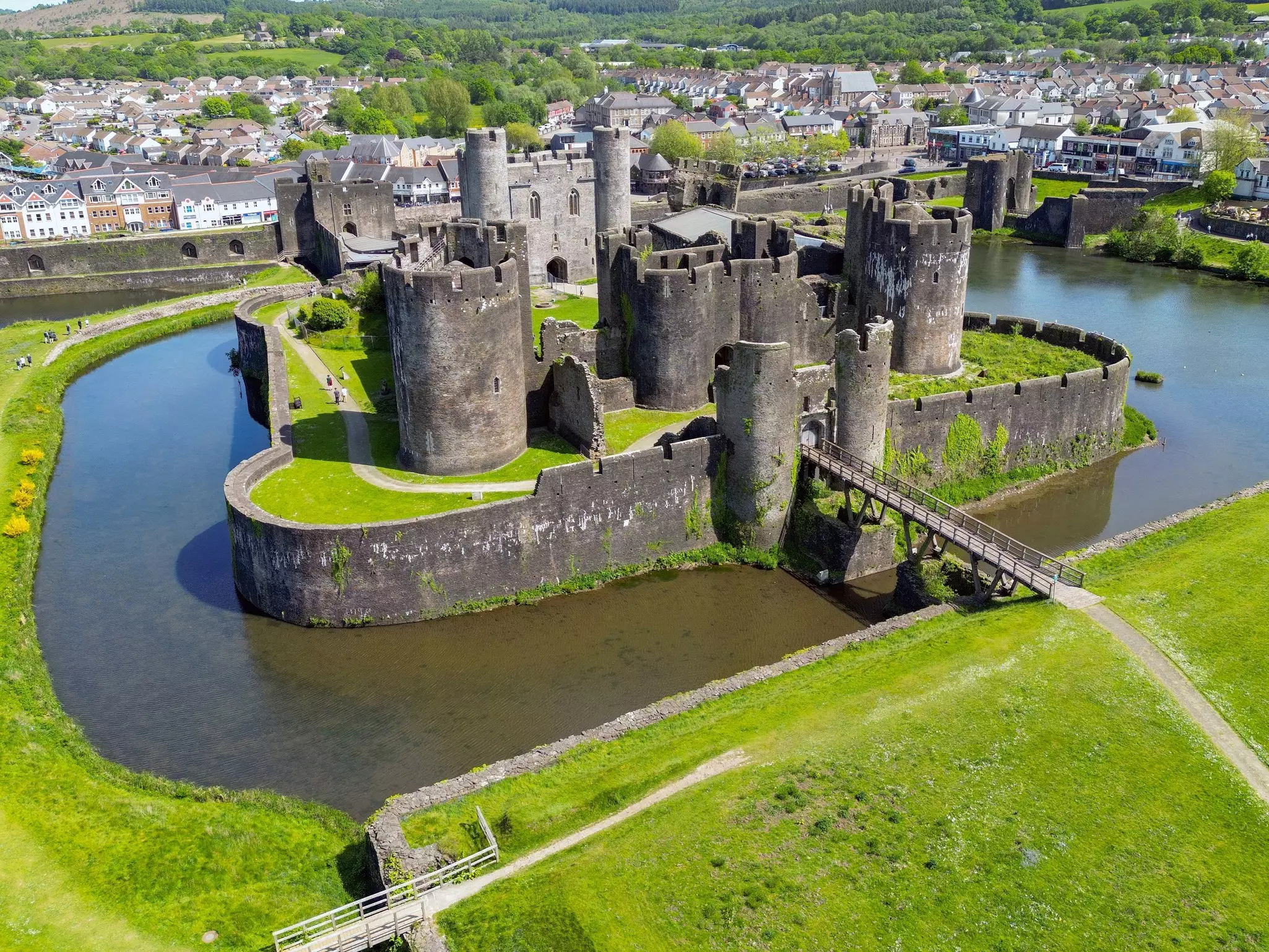 An aerial view of a largely intact medieval castle, with crenellated turrets, surrounded by a moat. A green lawn surrounds the moat, and a village is visible in the distance.