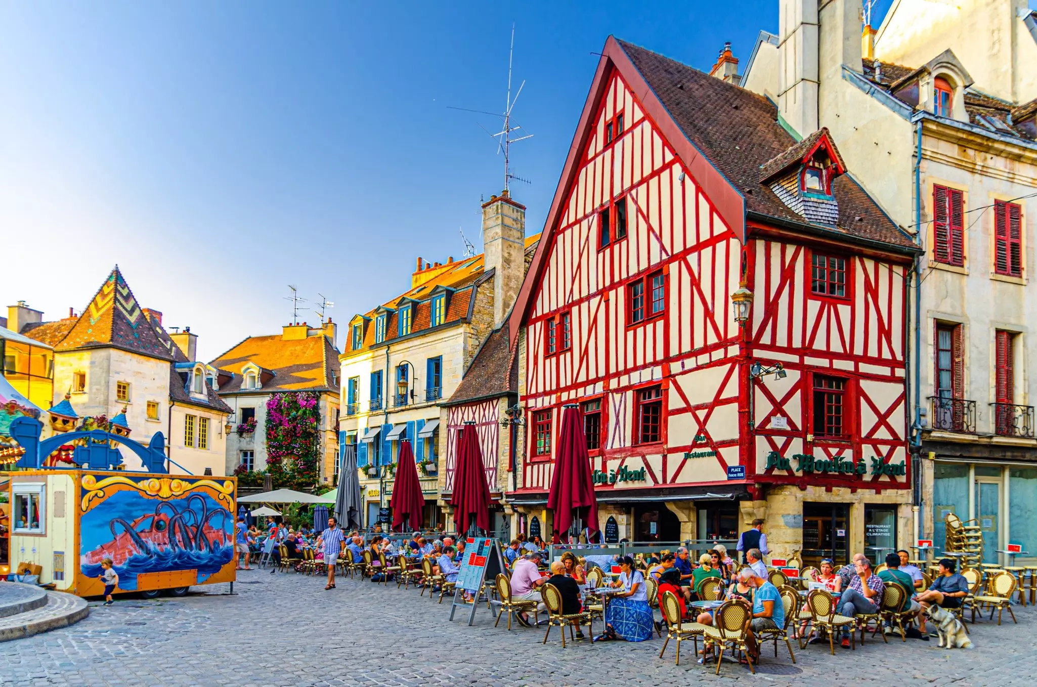 People sit at restaurant tables in a city square filled with historic buildings, one half-timbered.