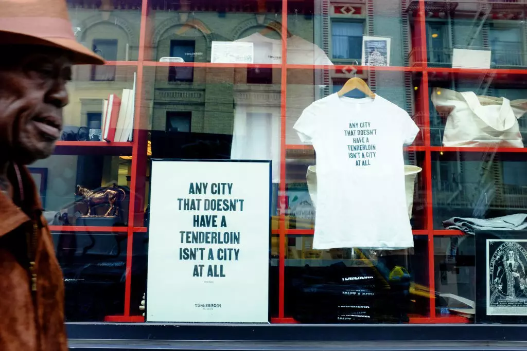 A man walks by a window with a poster and T-shirt hanging in the window that both read, "Any city that doesn't have a Tenderloin isn't a city at all.