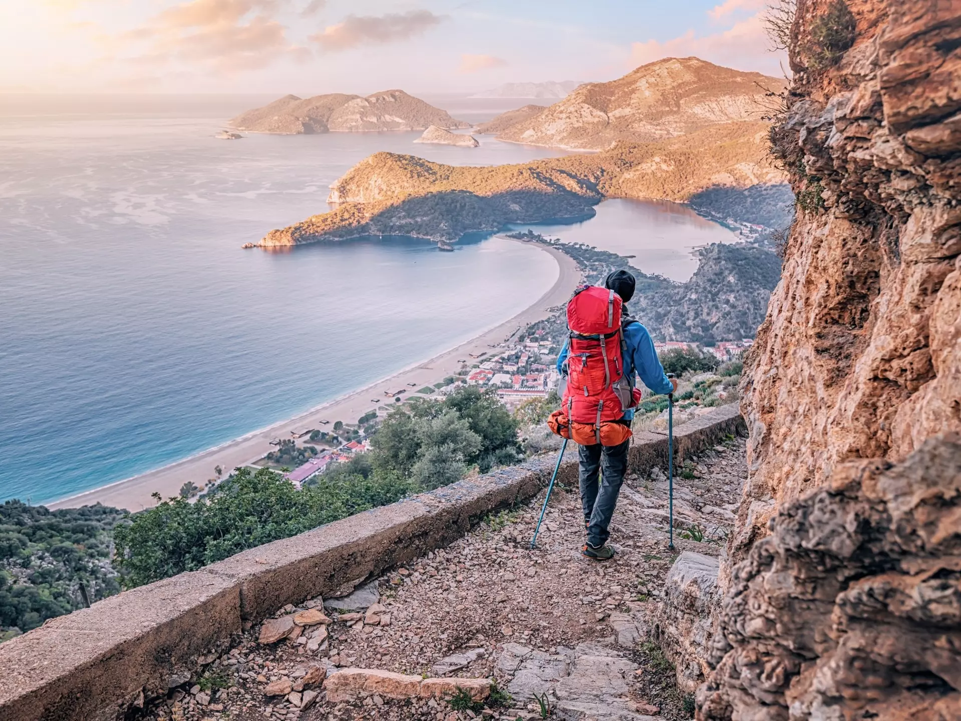 A hiker with a large backpack and walking poles navigates a rocky trail that hugs a coastal cliff. A view of the sea and other hilly capes is visible in the distance.