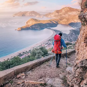 A hiker with a large backpack and walking poles navigates a rocky trail that hugs a coastal cliff. A view of the sea and other hilly capes is visible in the distance.