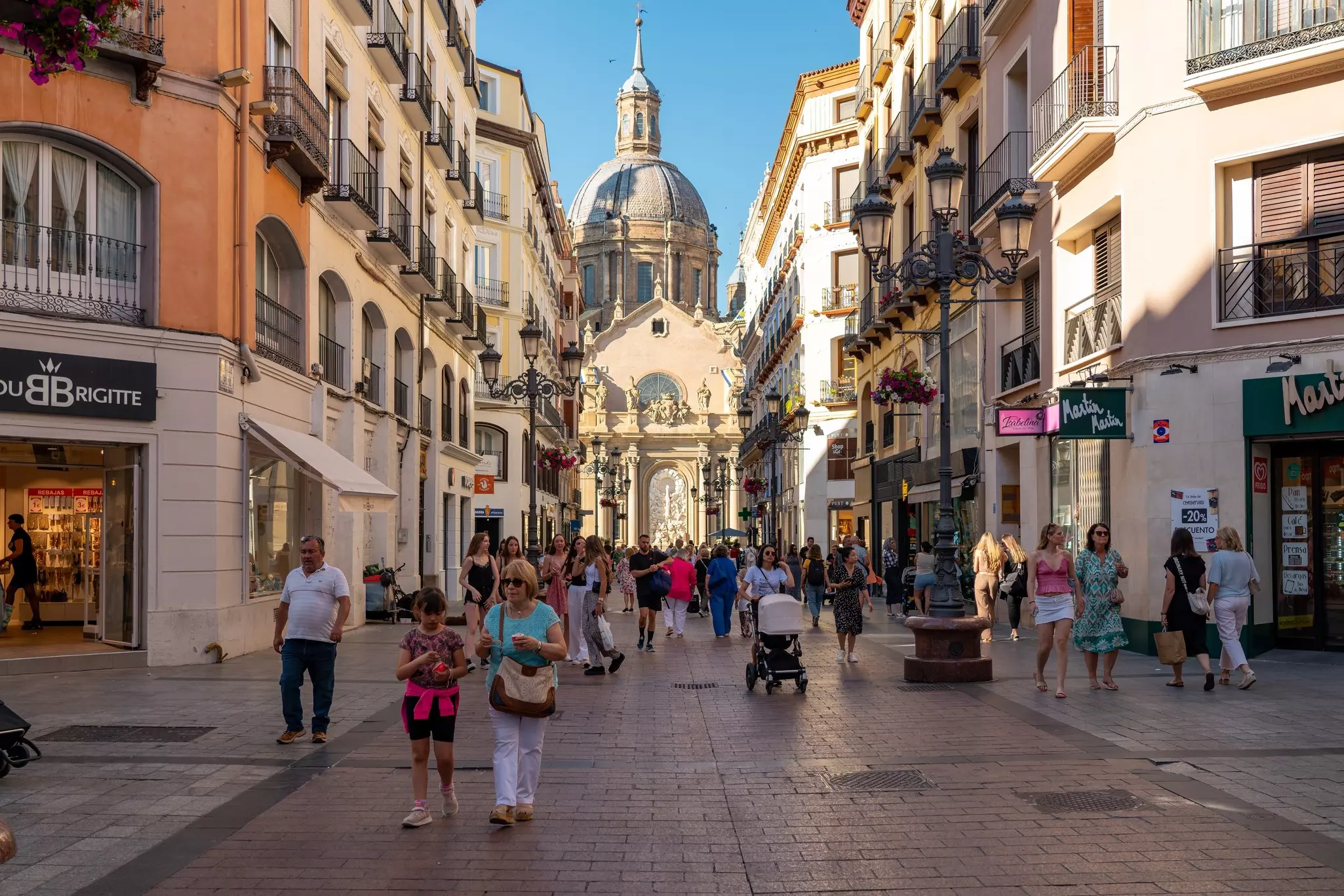 People wander down a pedestrianized street in a city near a large domed cathedral.