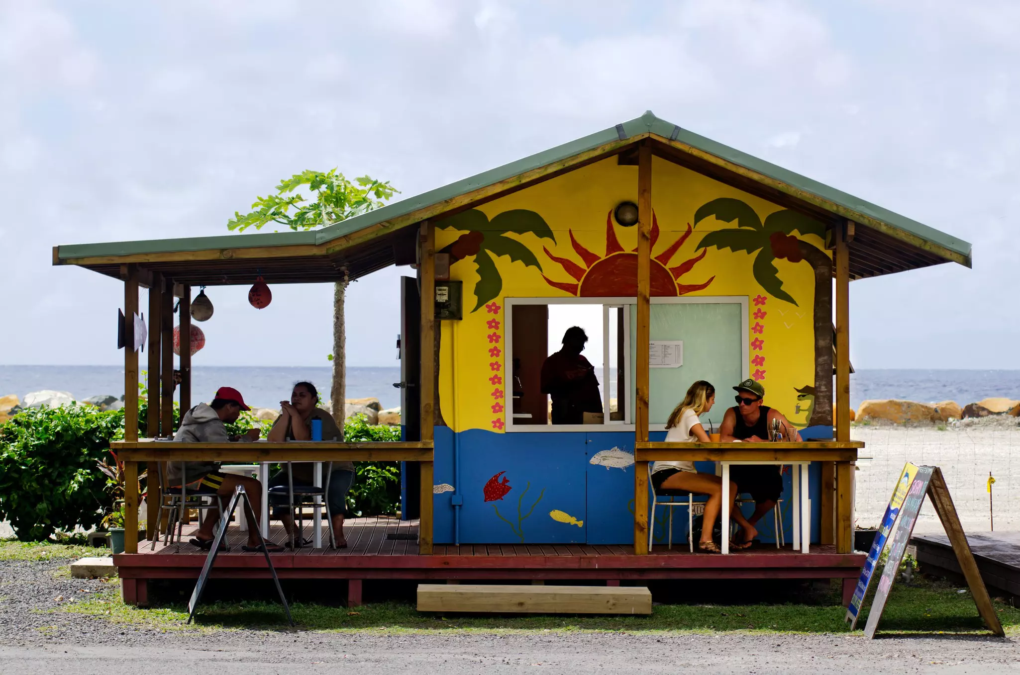A small restaurant occupies a brightly painted shack by the beach in a tropical destination. Patrons sit at tables enjoying their meals.