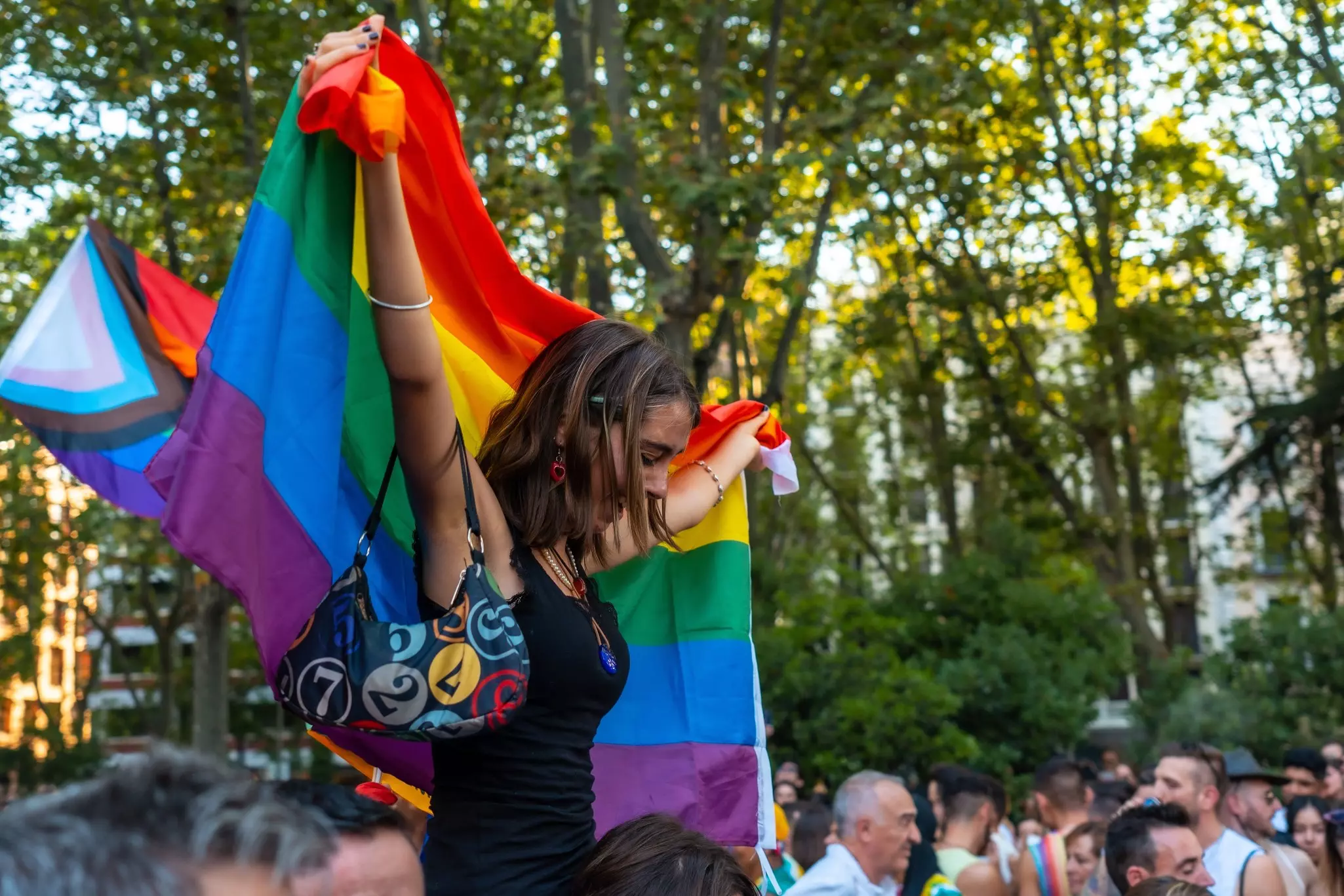 A woman sitting on someone's shoulders holds up a Pride flag during a parade
