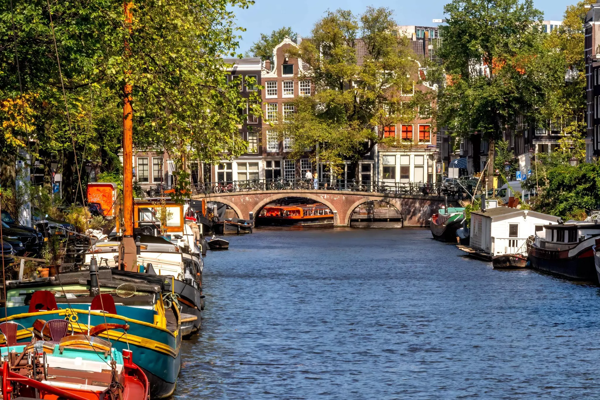 View of a canal on a sunny day, with canal boats on the left and right with a brick-sided bridge and brick buildings in the distance.