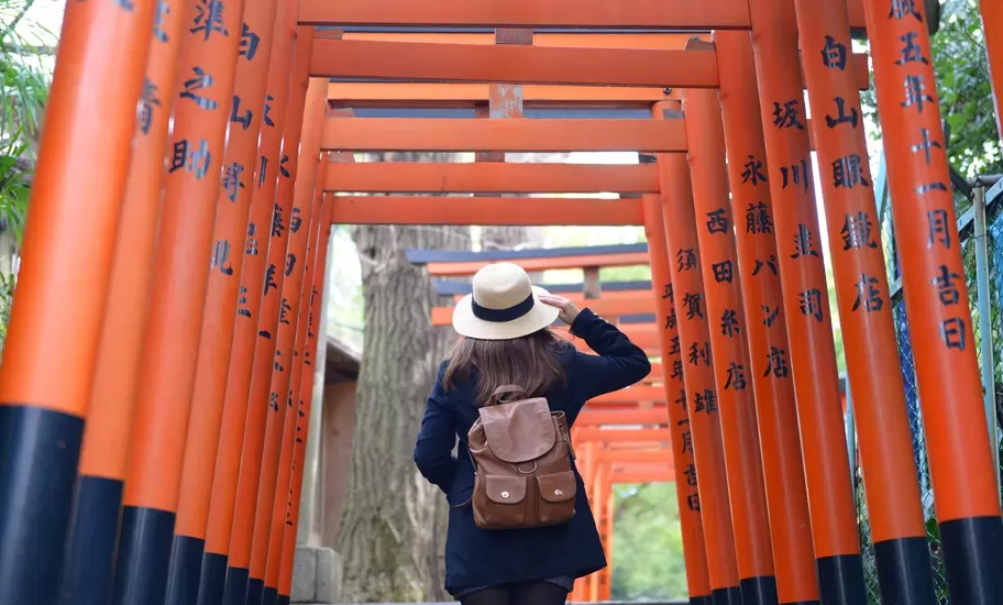 Woman in a straw hat with a leather backpack walks beneath a tunnel of vermilion torii gates inscribed with Japanese characters