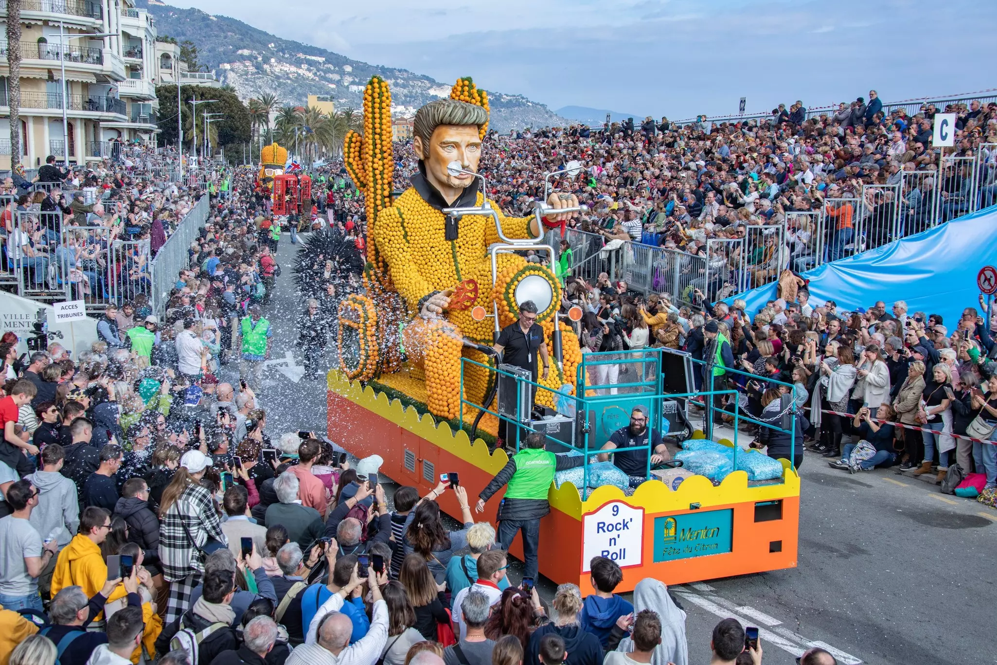 A street packed with revelers at a festival. A float in the shape of a man riding a motorcycle, made entirely of citrus fruits, parades down the street