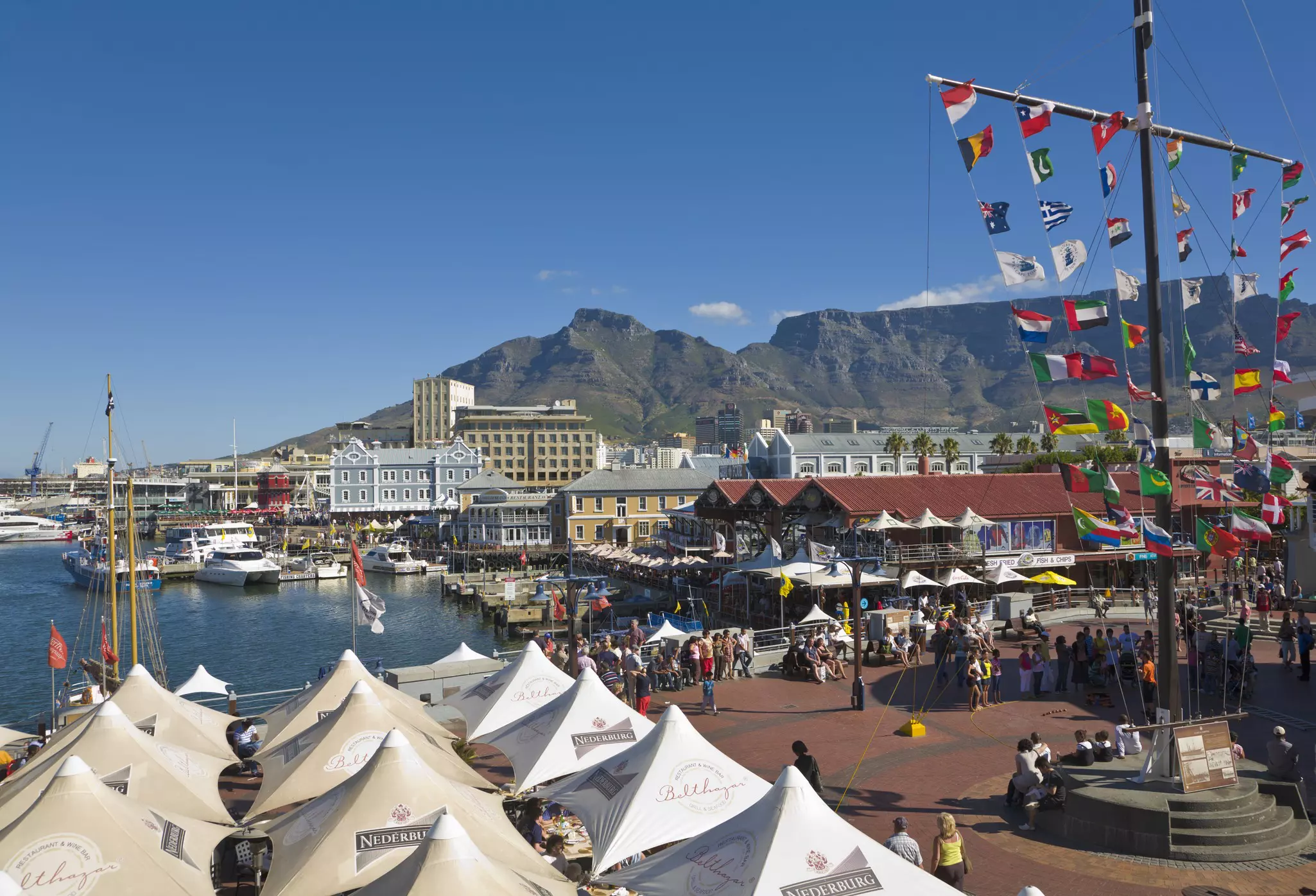 Tented awnings and commercial buildings in outdoor waterfront market with people walking and seated near a flagpole with multiple small, colorful flags on a sunny day. Boats are docked in the harbor in the distance.