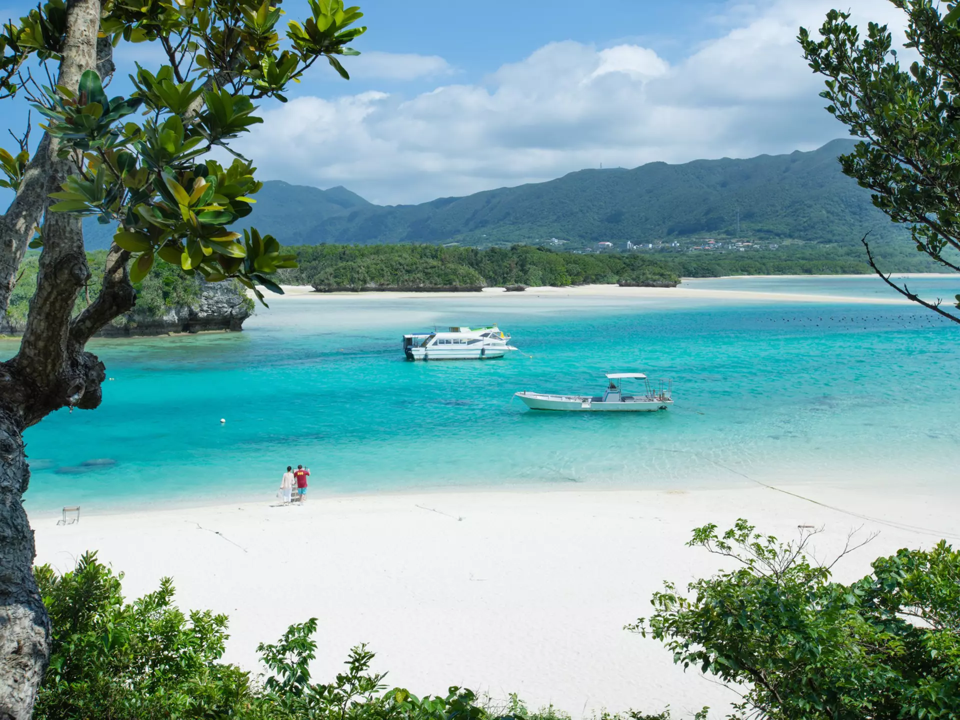 Two small boats moored in a peaceful turquoise lagoon at the edge of white sands on a tropical island. 