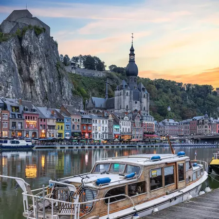 Pleasure boats moored along the River Meuse at dusk in Dinant, with diners at waterfront restaurants as evening light reflects along the water