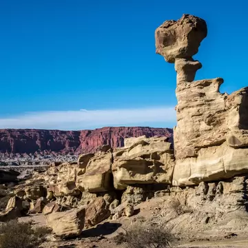 Sandstone hoodoo with a boulder perched on its narrow neck, red mesa cliffs and desert scrub behind, San Juan, Argentina