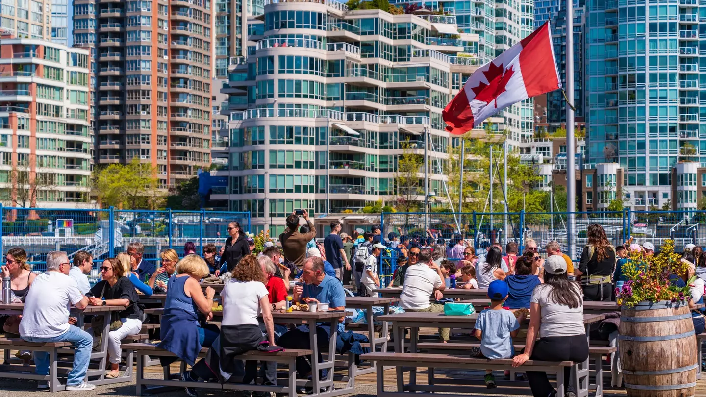People eat food at picnic benches in the sunshine in a high-rise city.
