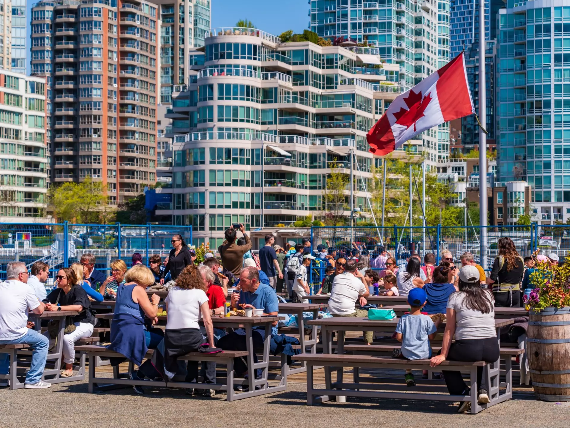 People eat food at picnic benches in the sunshine in a high-rise city.