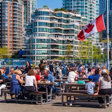 People eat food at picnic benches in the sunshine in a high-rise city.
