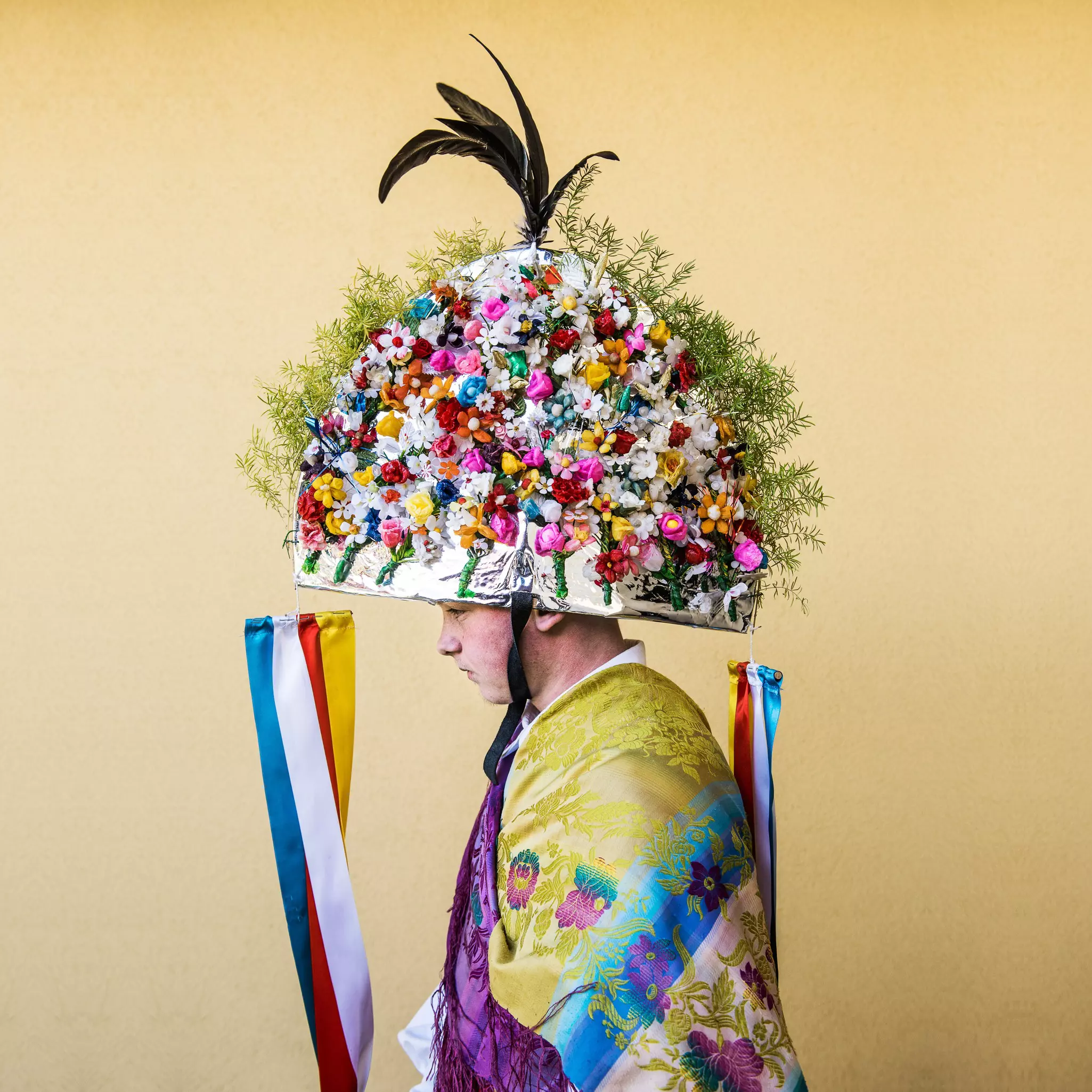 A person wears a large round headpiece covered with flowers and a patterned costume.