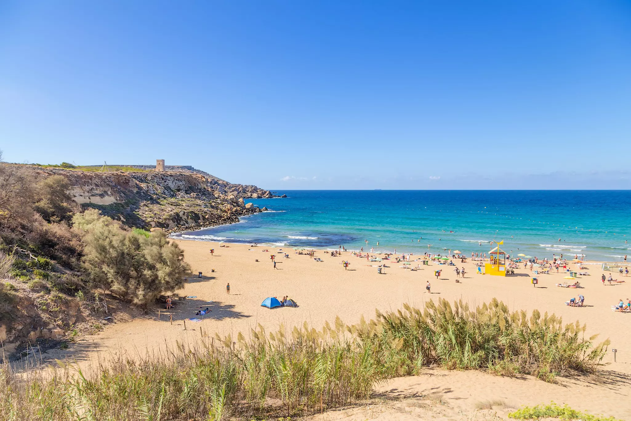 People relax on a gorgeous sandy beach with a tower on the top of a distant cliff