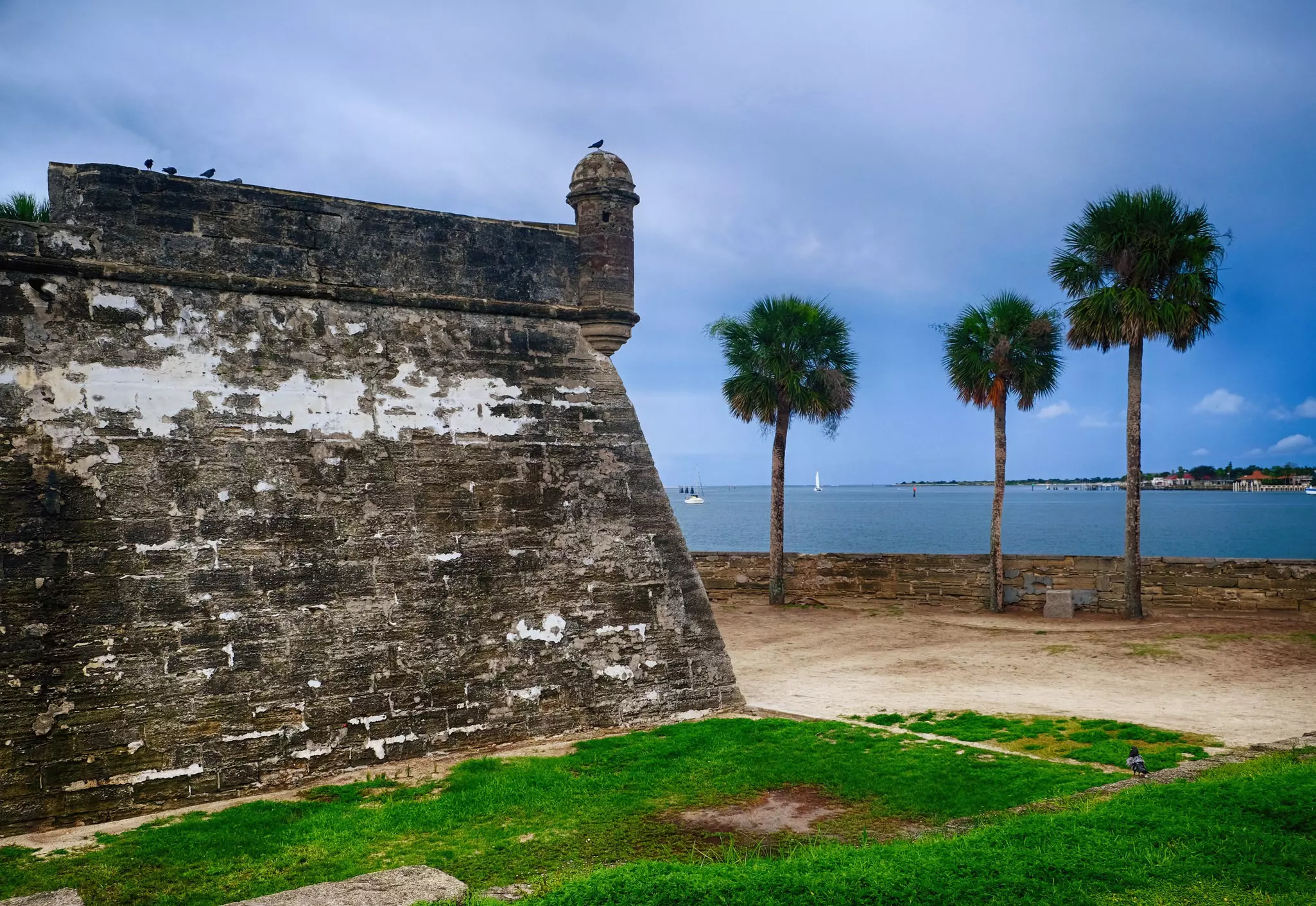 The exterior walls of a vast coastal fortress with adjacent palm trees and a stormy sky out at sea.