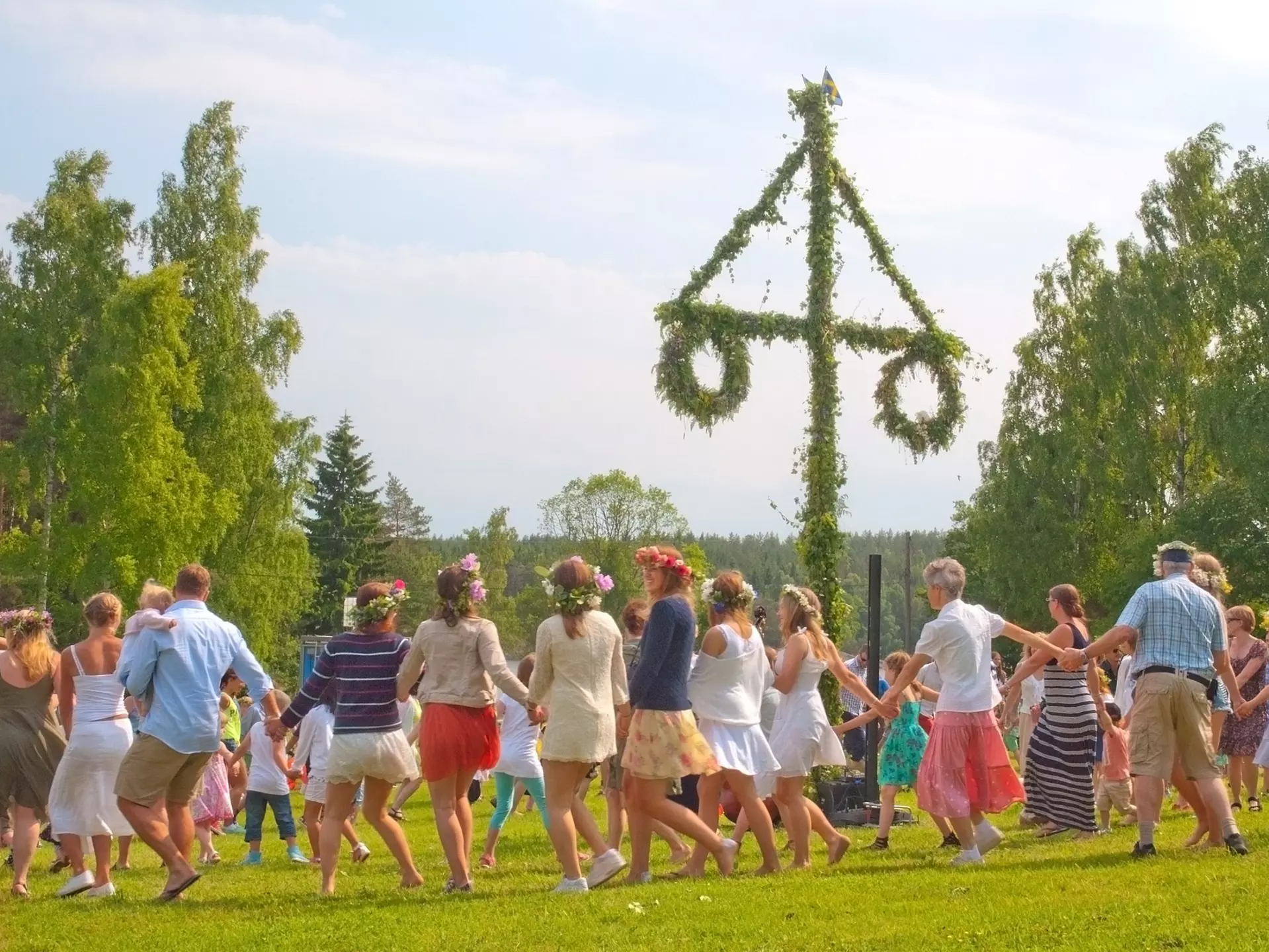 People holding hands and wearing flower crowns, dancing around a pole decorated with greenery.