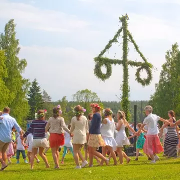 People holding hands and wearing flower crowns, dancing around a pole decorated with greenery.