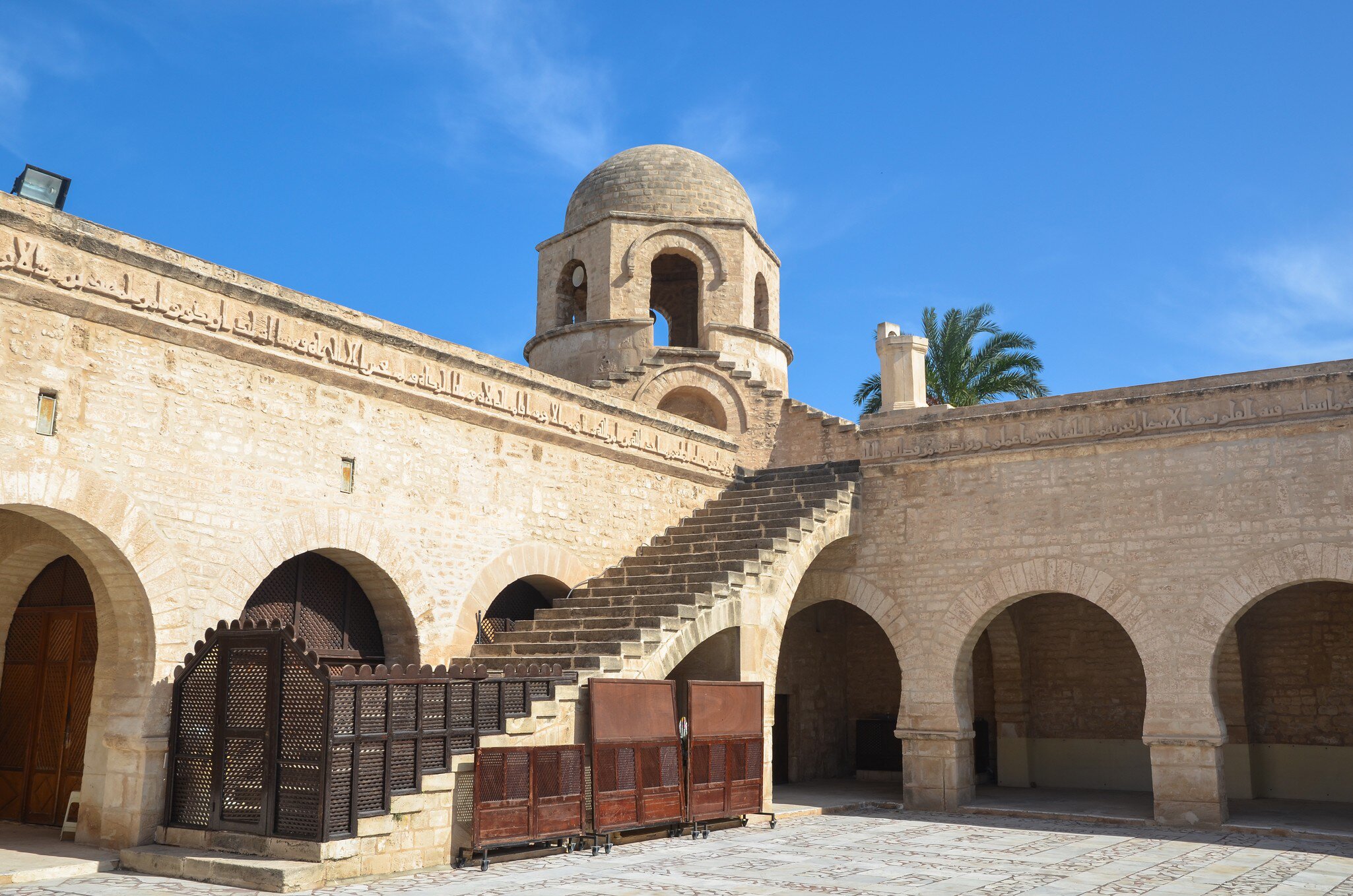 Pale stone arcades and carved inscription frieze, with an open staircase climbing to a small domed tower under blue sky