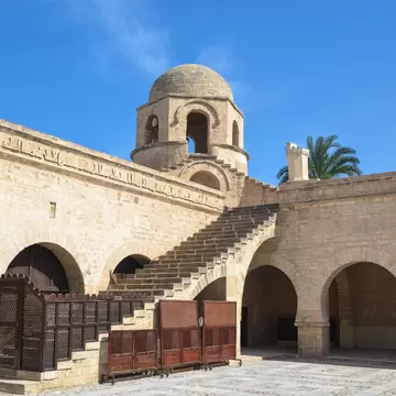 Pale stone arcades and carved inscription frieze, with an open staircase climbing to a small domed tower under blue sky