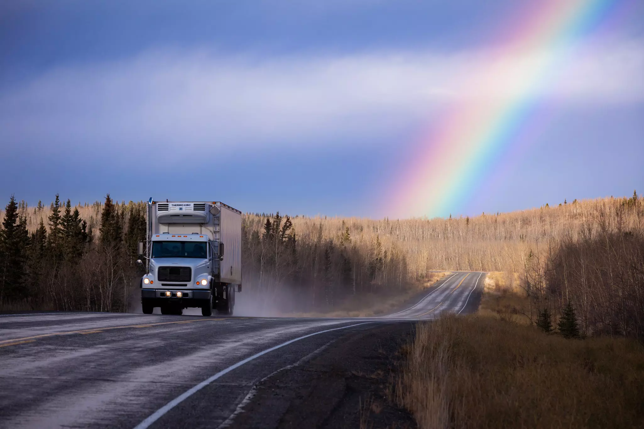 A truck drives along a highway through a forest, with a large rainbow pictured in the distance.