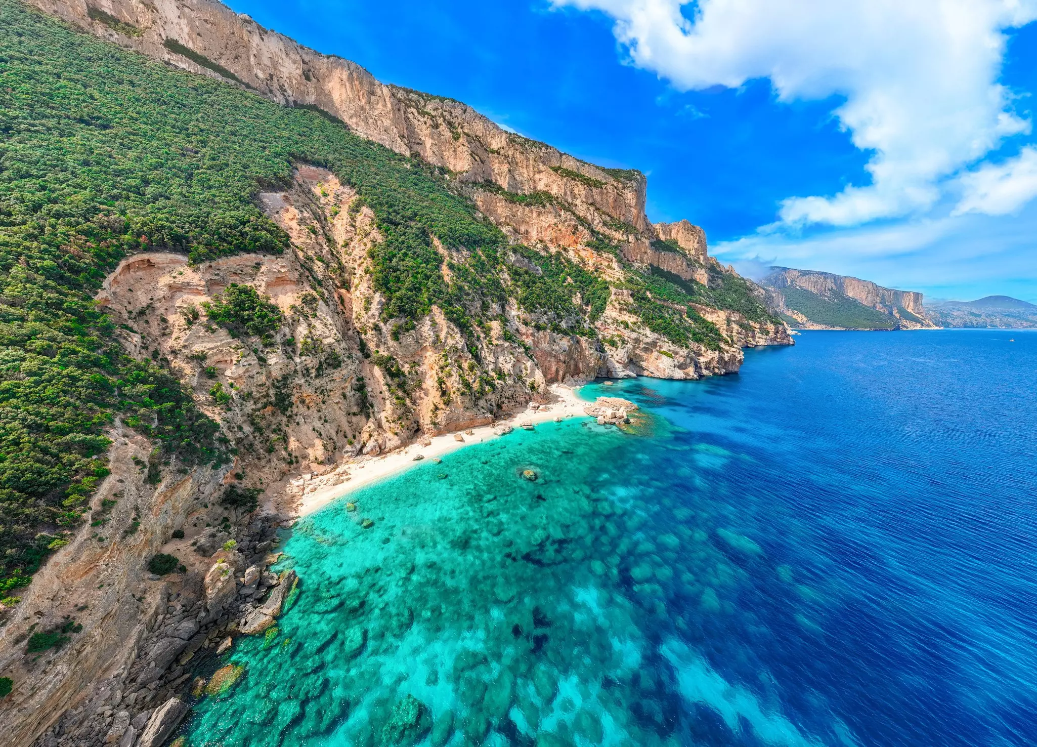 Aerial view of turquoise sea with rocky cliffs to the left and in the distance on a sunny day.