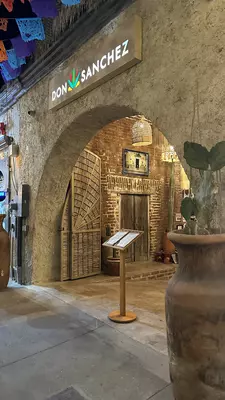 Front of museum with colorful flags hanging above it.  Colorful painting of a woman can be seen through the doorway. 