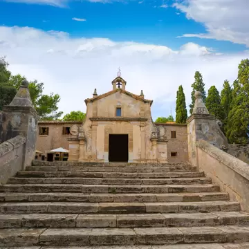 Small weathered chapel with a bell gable at the top of a broad stone stairway in Pollença, flanked by cypress trees