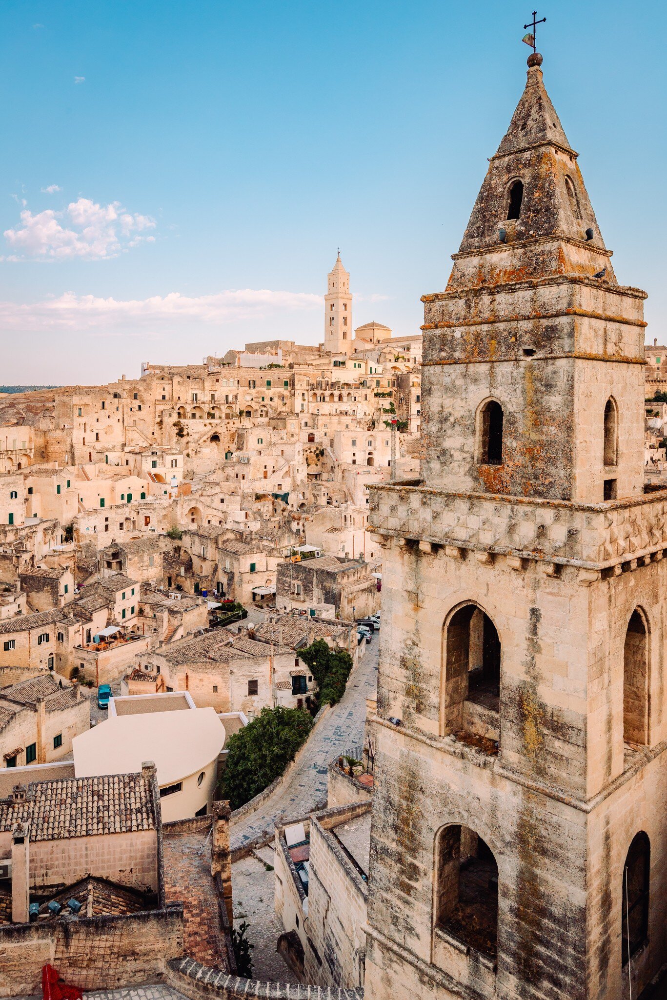 Weathered stone bell tower in the foreground above a hillside of pale stone cave houses, with a cathedral tower on the ridge.