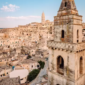 Weathered stone bell tower in the foreground above a hillside of pale stone cave houses, with a cathedral tower on the ridge.