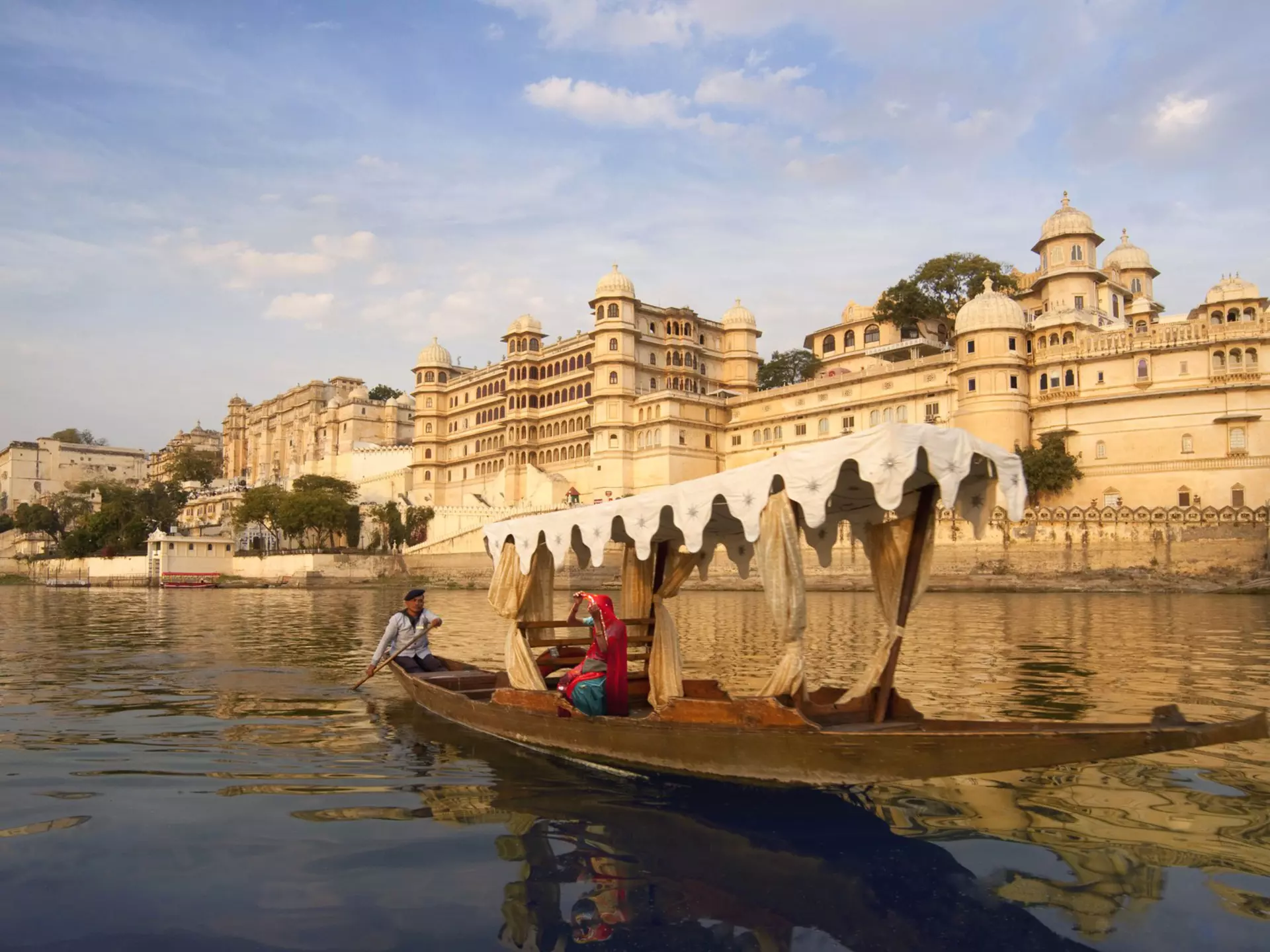 A person rowing a boat with an awning past a tan stone palace.