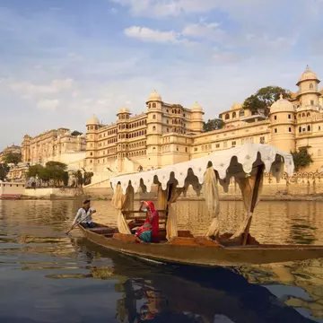 A person rowing a boat with an awning past a tan stone palace.