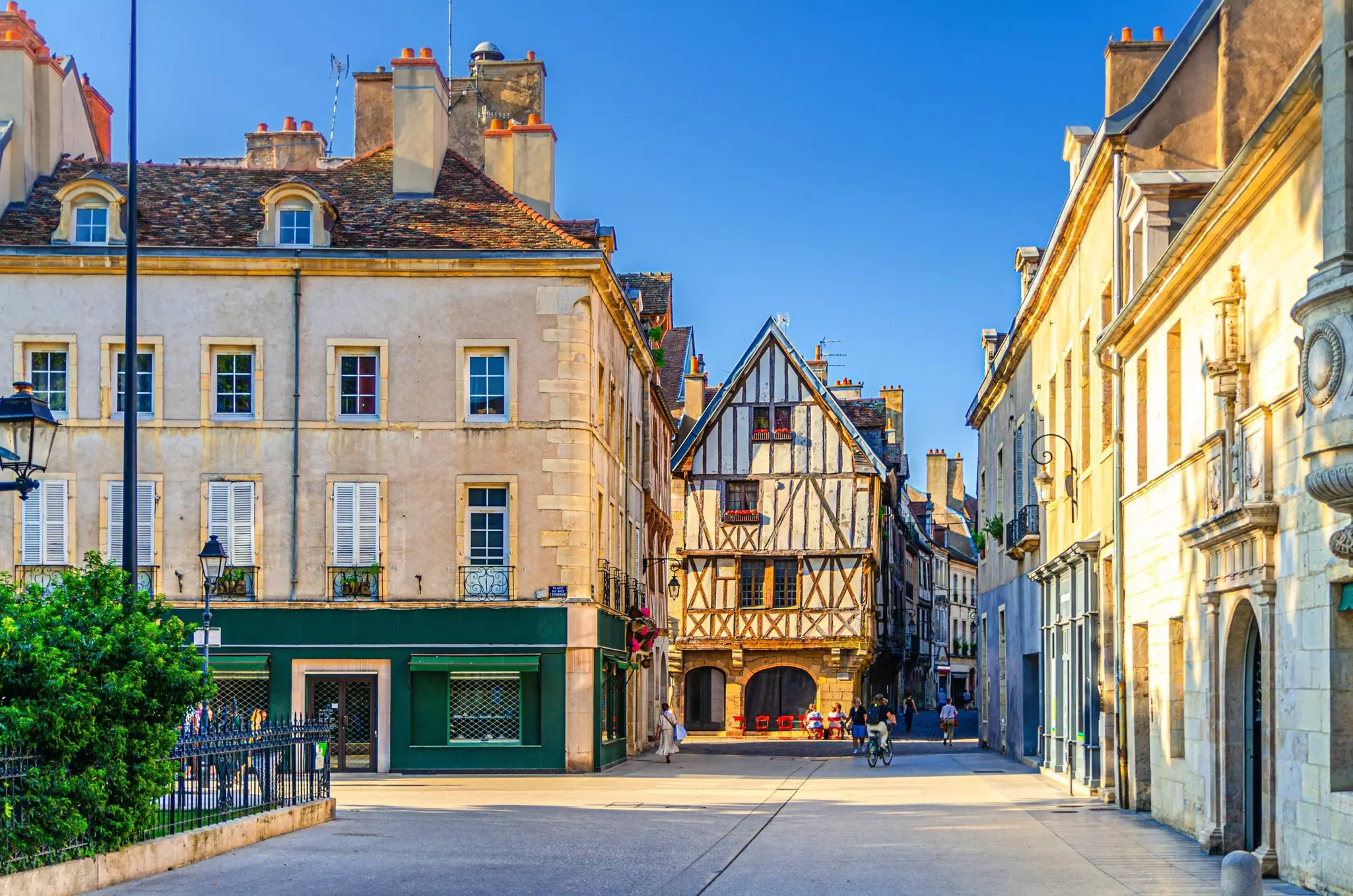 A road off a city square leads to a half-timbered building on a sunny day. People are walking and cycling nearby.