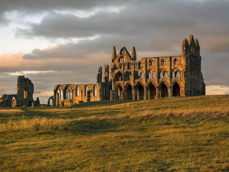 The ruins of a Gothic abbey with many crumbling arches on the top of a windswept headland bathed in sunlight.