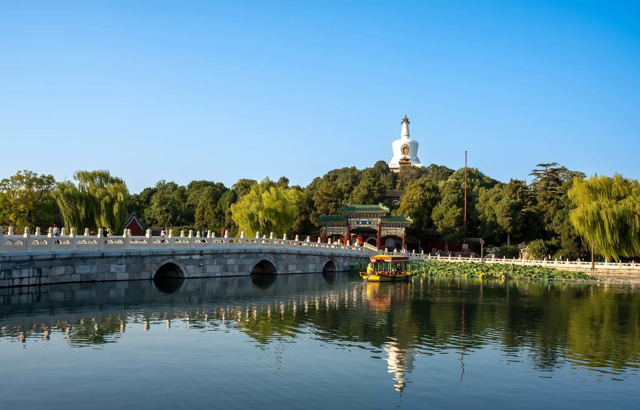 Wide shot of water with cement bridge and yellow boat with buildings and green trees in the background.