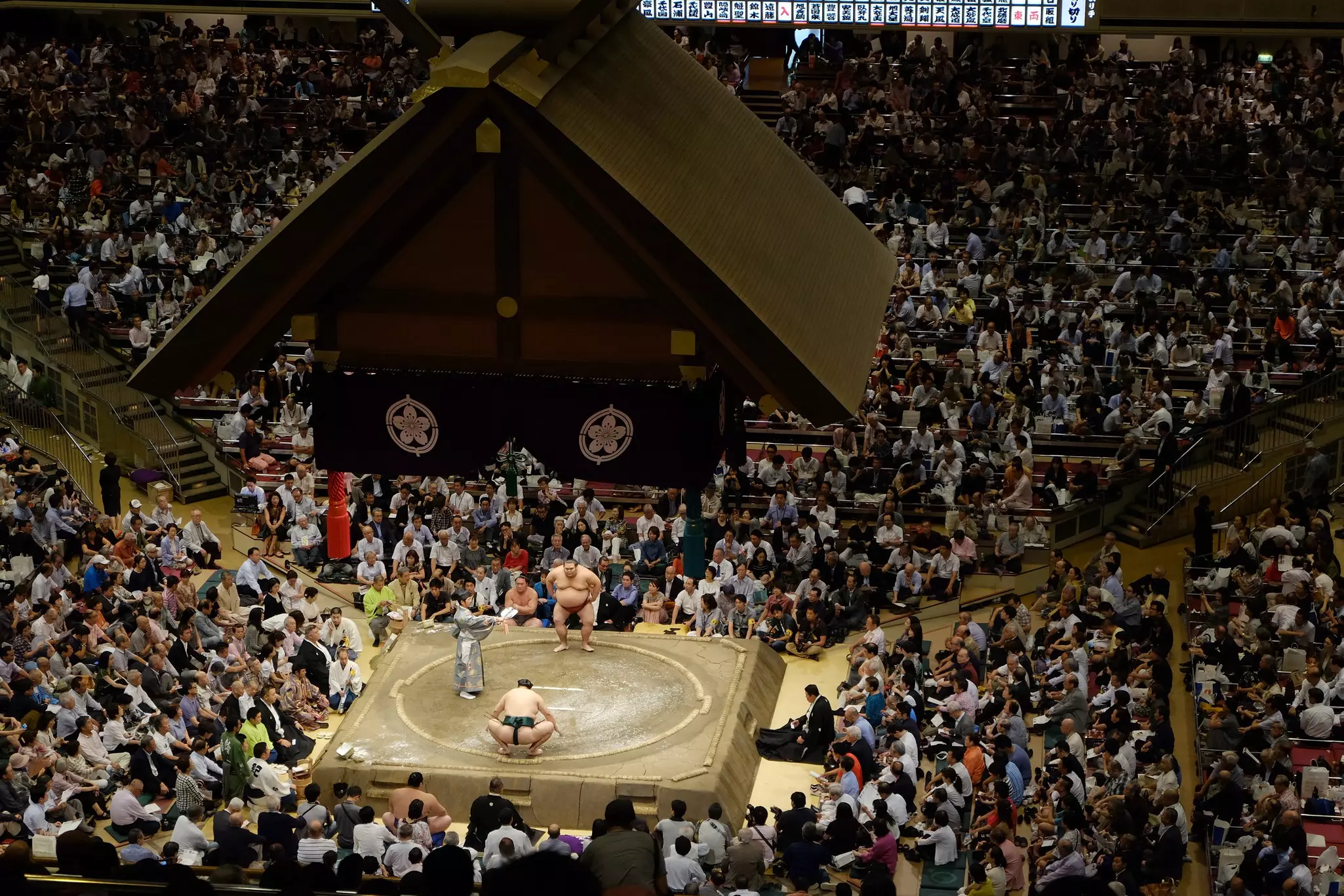 A wide view of an arena, with tiers of seats leading to a small wrestling ring capped by a traditional canopy.