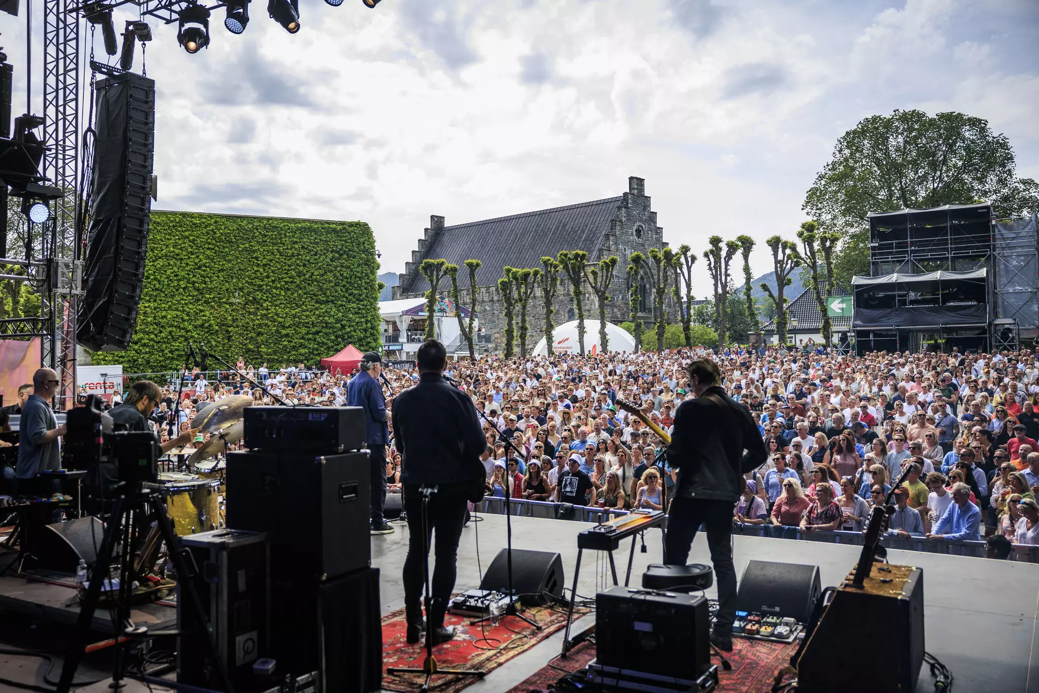 A band performs, pictured from behind, in front of a large crowd of spectators. Historic buildings are seen behind the crowd.