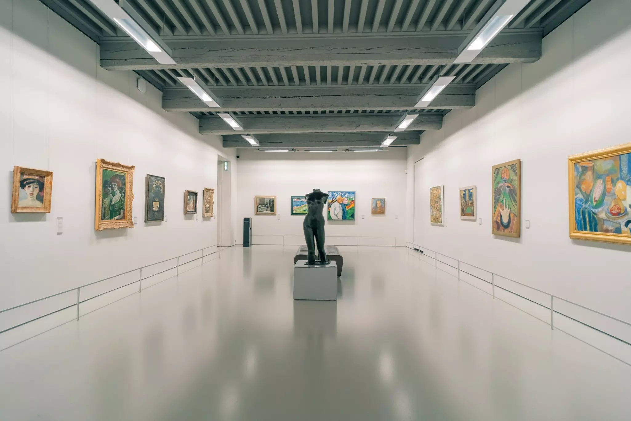 A sculpture stands in the center of a museum gallery whose walls are hung with paintings.