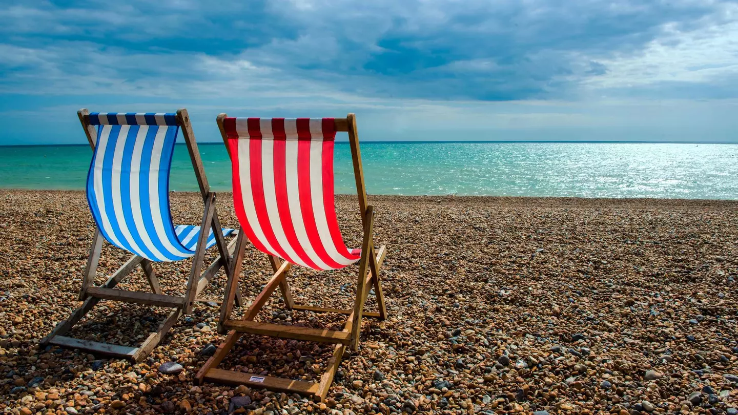 Two empty deck chairs, one with red and white stripes, the other with blue and white stripes, are arranged to look out towards the sea on Brighton beach. The fabric is billowing in the wind.