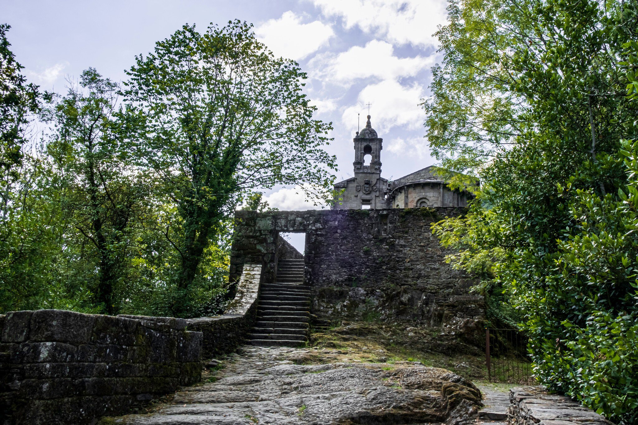 View of stone steps leading to Mosteiro de Caaveiro in Spain, framed by green trees.